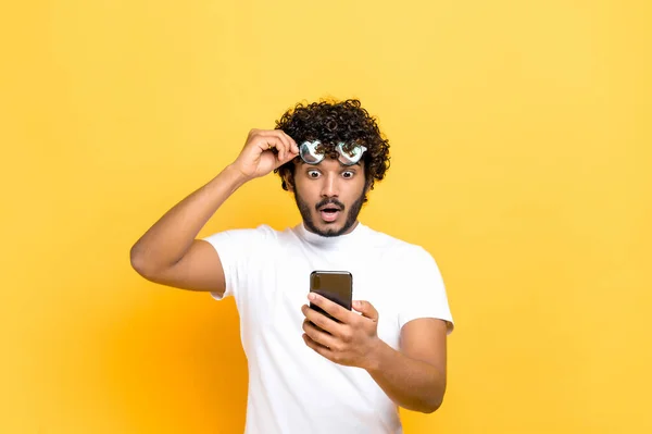 Amazed shocked unshaven Indian or Arabian guy holding smartphone in his hand, looking at the camera in surprise with his glasses raised, shocked facial expression, isolated orange background — Stock Photo, Image