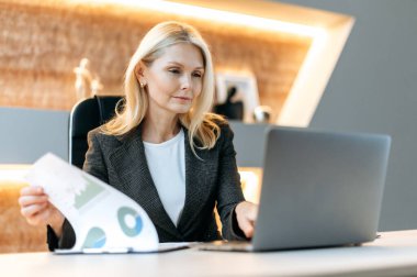 Kafkasyalı olgun, başarılı, başarılı, zeki iş kadını, finansal analist, yönetici, dizüstü bilgisayarda oturuyor, belgeleri okuyor, şemaları analiz ediyor ve finansal strateji planlıyor.