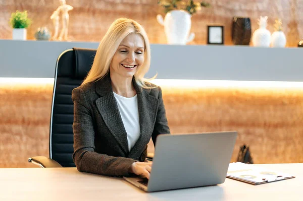 Rijpe schattige gelukkige succesvolle blanke zakenvrouw, top manager, bedrijf makelaar, in formele pak, zitten aan tafel in kantoor, met behulp van laptop, sms 'en met klant of collega' s, glimlachend vriendelijk — Stockfoto