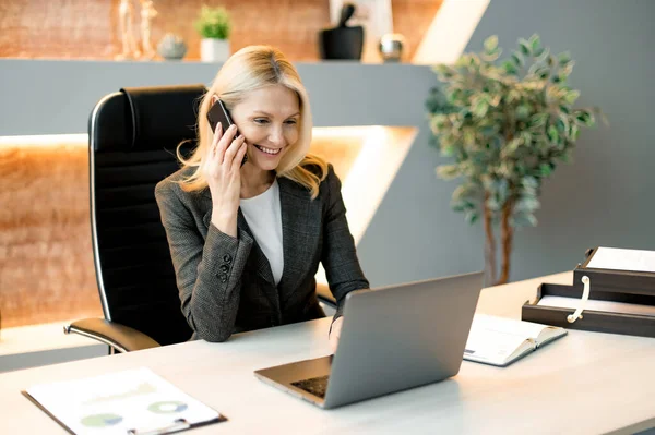 Drukke vriendelijke succesvolle intelligente blanke zakenvrouw, freelancer of top manager zitten aan tafel in het kantoor met laptop, praten met de klant of medewerkers op mobiele telefoon, glimlachen — Stockfoto
