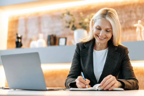 Gelukkige slimme blanke zakenvrouw, tom manager, ceo, zitten aan een tafel in een modern kantoor, met behulp van een laptop, nadenken over zakelijke strategieën en financiële mogelijkheden, het maken van notities, glimlachen — Stockfoto
