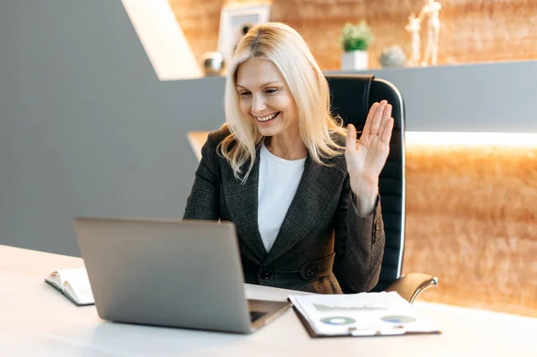 Positive beautiful influential Caucasian mature woman, top company manager, sits in office using laptop, talking online with client or employees, discussing project, holds consultation, waving hand — Stock Photo, Image