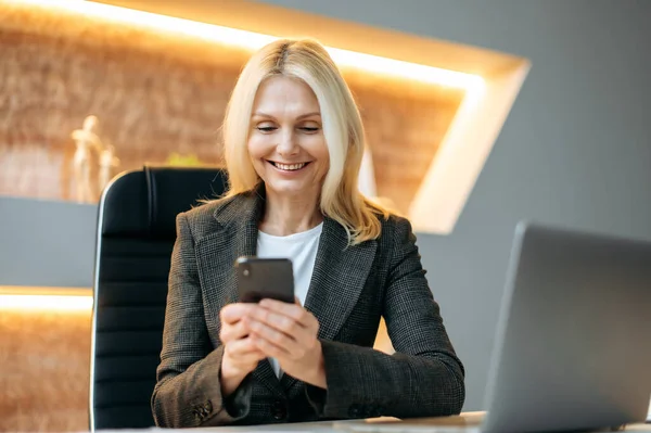 Friendly happy cute mature caucasian businesswoman or manager using smartphone, sitting at table in office, texting with client, friends or colleagues, got good news, writing sms, smiling — Stock Photo, Image