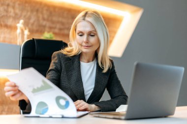 Odaklanmış zeki Kafkasyalı olgun iş kadını, finans analisti, en iyi yönetici, ofisinde oturmuş, finansal çizelgeler okuyor, analiz ediyor ve finansal strateji planlıyor.