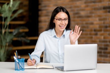 Şirin, kendinden emin, zeki, beyaz iş kadını, serbest çalışan, gözlüklü ya da menajer, ofiste masada oturuyor, iş arkadaşlarıyla veya müşterileriyle video iletişimi kuruyor, el sallıyor, gülümsüyor.