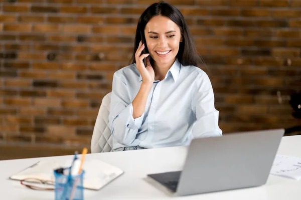 Joyeux amical caucasien brunette, femme d'affaires, spécialiste de l'informatique, gestionnaire, assis au bureau à l'ordinateur portable, parlant au téléphone tout en travaillant avec des amis ou un client, souriant amical, regardant l'écran — Photo