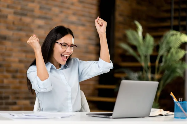 Happy joyful successful smart emotional caucasian business woman, broker, freelancer wearing glasses, sitting in office with laptop joined good deal, victory, good news, gesturing with fists smile — Stok Foto