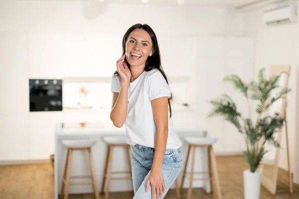 Beautiful happy cheerful Caucasian young adult brunette woman, in casual stylish clothes, stands in modern living room at home, looking at camera, smiling friendly, looks happy — Stockfoto