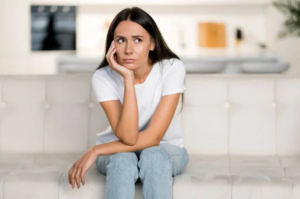 Disappointed unhappy caucasian young woman, sitting on the sofa in the living room, she has personal problems, failure at work, depression, bad mood, sad girl is lonely at home