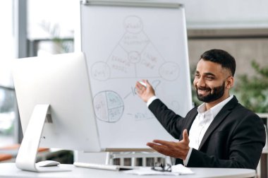 Distant meeting. Confident successful influential Indian business coach or businessman in formal suit sit in office, conducting online seminar for colleagues or students showing diagrams on whiteboard