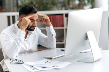 Upset young adult Indian, company owner, manager or office worker with beard, sits at his desk in modern office with closed eyes, rubs his head, feels stressed and tired from overtime work, needs rest