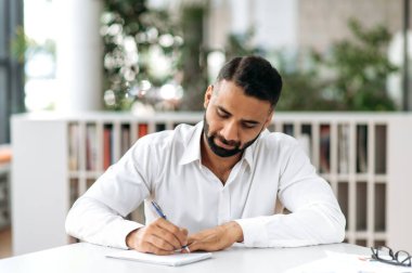 Concentrated serious smart confident Indian business man, manager or freelancer, wearing white shirt, sitting at table in modern office and taking notes, listening online webinar, brainstorm