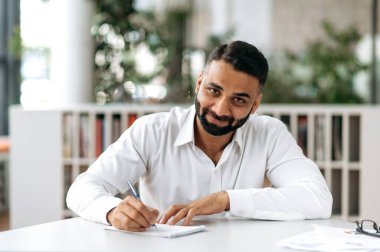 Positive successful Indian business person, manager or freelancer, wearing white shirt, sitting at table in modern office and taking notes, listening online webinar, brainstorm. E-learning concept