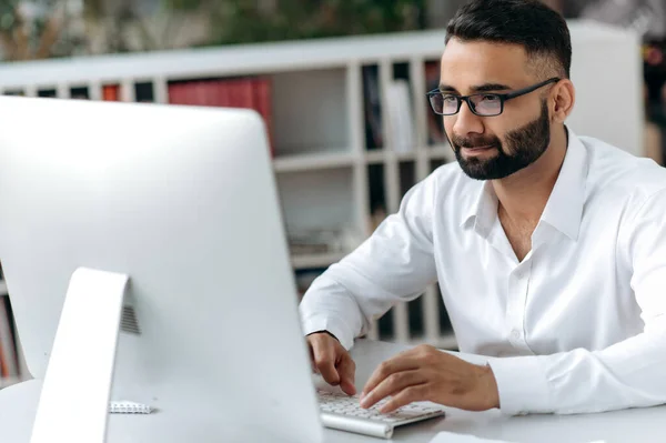 Successful Indian businessman, manager or office worker with beard and glasses, sitting at desk in modern office, wearing white shirt, using computer, texting with colleagues or client, analyzing — Stok fotoğraf