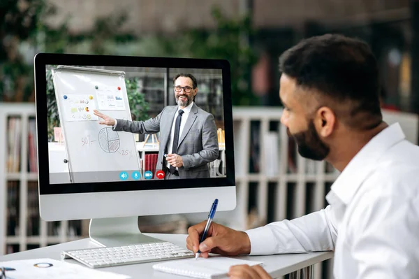 Online business video conference. Concentrated top manager or business owner sitting at desk in office, wearing white shirt, listening online business training, watching coach video, taking notes — 스톡 사진