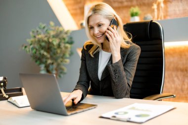 Mature happy joyful confident caucasian blonde business lady, broker or manager, sitting at table in a modern office, working with laptop, talking to client or employees on smartphone, smiling