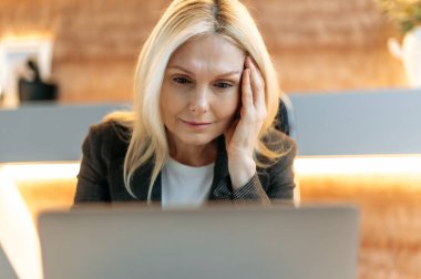 Smart focused exhausted mature woman, business woman or broker, in formal suit, tired of office work, experiencing stress and headache, massaging her temple, needs rest, looking languidly at screen