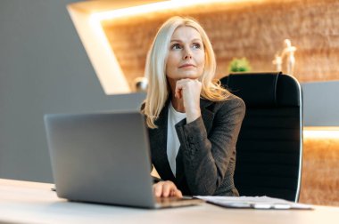 Influential successful smart blonde caucasian mature business woman, financial analyst, freelancer, sitting at table, taking break from work, looking thoughtfully aside, dreaming about vacation