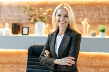 Business portrait happy successful smart caucasian mature business woman, top manager, broker, standing in modern office, wearing formal stylish suit, looking at camera, arms crossed, smiling friendly