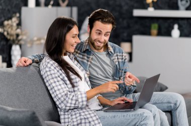 Positive caucasian couple using laptop while resting on couch at living room at home, spending time together, browsing social media, internet, watches videos, messaging with friends, smiling