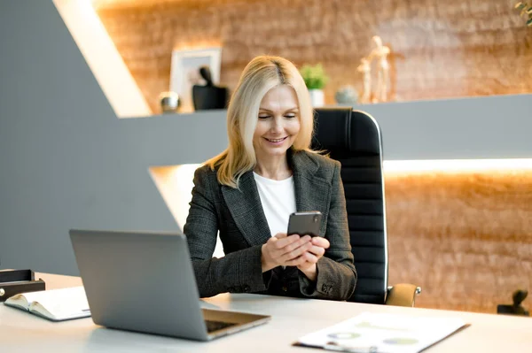 Joyful successful attractive influential mature caucasian businesswoman or top manager using cellphone while sitting at desk in office, texting client, friends or colleagues, got good news, messaging — Stock Photo, Image