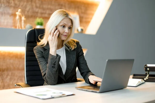 Focused smart surprised successful influential caucasian businesswoman, freelancer or top manager sitting at table in an office using laptop, talking by cellphone, browsing internet or e-documents — Stockfoto