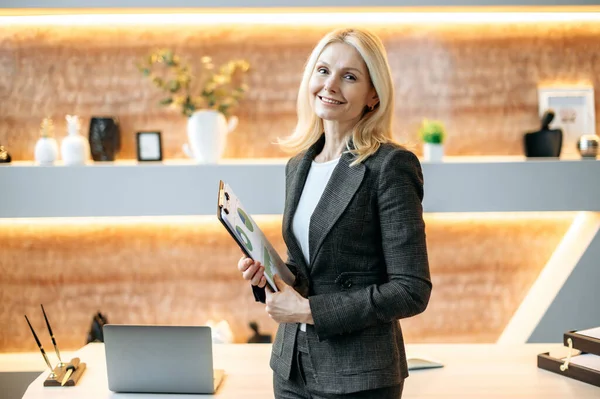 Happy successful mature caucasian businesswoman or corporate top manager, standing near work desk in modern office, dressed in stylish suit, holds business documents, looking at camera, smile friendly — Stockfoto
