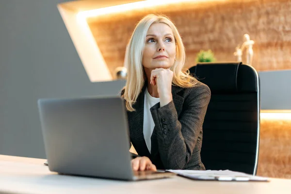 Influential successful smart blonde caucasian mature business woman, financial analyst, freelancer, sitting at table, taking break from work, looking thoughtfully aside, dreaming about vacation — Stok fotoğraf