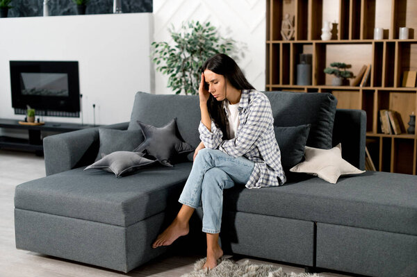 Sad caucasian brunette woman in stress, sitting on the sofa alone, in casual clothes, experiencing headache, migraine, massaging her temple, closing her eyes, need rest and treatment