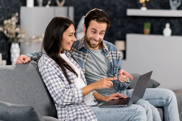 Positive caucasian couple using laptop while resting on couch at living room at home, spending time together, browsing social media, internet, watches videos, messaging with friends, smiling — Foto de Stock