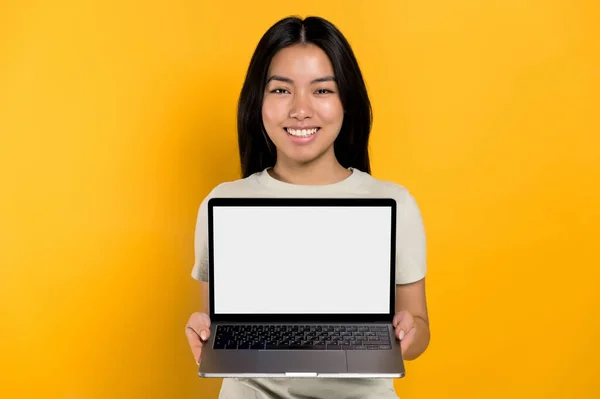 Happy chinese positive young woman in casual t-shirt holding open laptop with blank white screen, stands on isolated orange background, looks at camera, mockup for your advertisement, copy space — Stockfoto