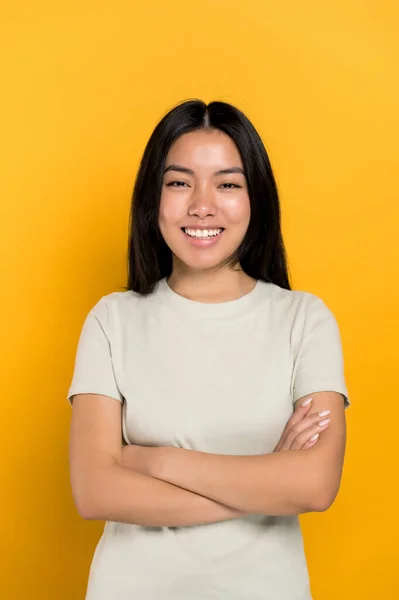 Portrait of a pretty, lovely positive long-haired brunette Asian modern girl in casual t-shirt, looking directly at the camera, smiles friendly,standing on isolated orange background with arms crossed — стокове фото