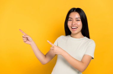 Happy pretty brunette millennial Chinese young woman, points with hands to empty space, excited looks at camera, smiling friendly, stands on isolated orange background in casual t-shirt