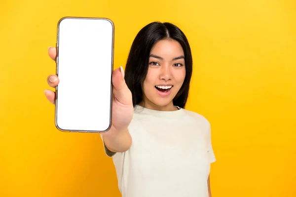 Lovely excited chinese positive brunette young woman in casual t-shirt showing modern smart phone with blank white screen, standing on isolated yellow background, smiling. Mockup, copy space — Stockfoto