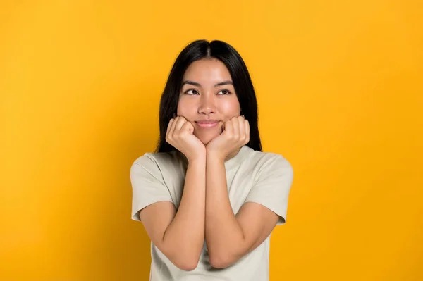 An dreaming positive millennial Asian brunette creative girl, dreaming about something, looks at empty space, thinking, make choice, standing on isolated orange background, smiling. Copy space — 스톡 사진