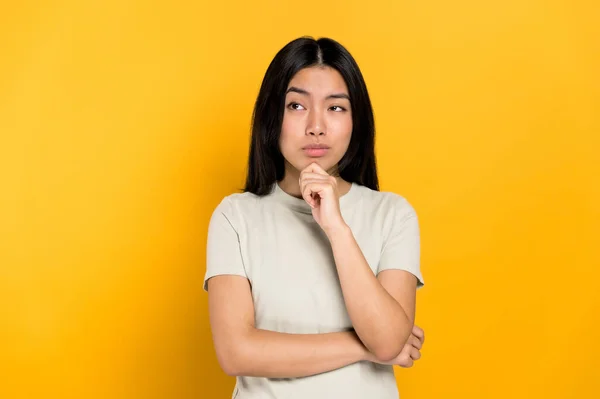 Pensive millennial smart Asian brunette young woman, minded, thinking, creative girl, look up empty space, developing idea, make choice, standing isolated orange background. Copy space — стокове фото
