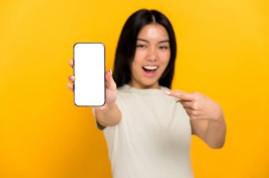 Defocused chinese positive young woman in casual t-shirt showing modern cell phone with blank white screen and points a finger at him, stands on isolated yellow background, mockup, copy space