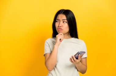 Unsure, Asian or Chinese young long-haired brunette woman, hold a cell phone, puzzled look empty space, thinking, planning, dreaming, standing on isolated orange background