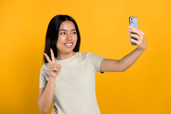 Lovely happy excited chinese brunette girl in casual t-shirt, doing selfie shot on smartphone and smiles into the camera, standing on isolated orange background — ストック写真