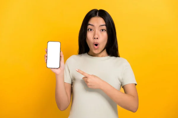 Shocked amazed chinese brunette girl in casual t-shirt showing modern smartphone with blank white screen and points a finger at him, stands on isolated orange background, mockup, copy space — Stockfoto