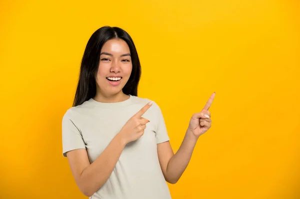 Happy lovely millennial brunette long-haired Asian young woman, points with hands to empty space, looks at camera, smiling friendly, stands on isolated background in casual t-shirt — Zdjęcie stockowe