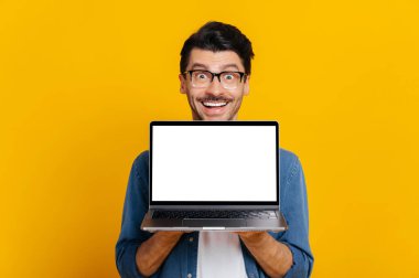 Happy caucasian positive man in casual modern wear, holding open laptop with blank white screen, stands on isolated orange background, looks at camera, mockup for your advertisement, copy space