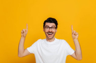 Positive caucasian young man in white basic t-shirt and glasses amazed looks at the camera and points fingers up at empty space, stands on isolated orange color background
