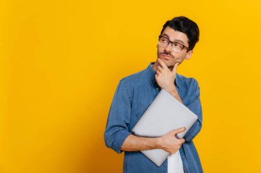 Puzzled smart guy in glasses and in a denim shirt, holding a laptop at hand, looks thoughtfully towards empty space aside touching his chin, smiling, stands on isolated orange background, copy space