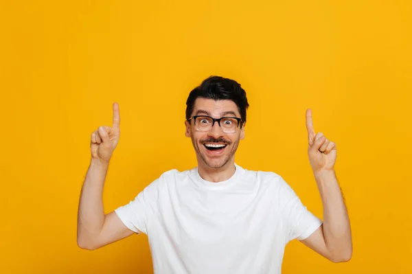 Positive caucasian young man in white basic t-shirt and glasses amazed looks at the camera and points fingers up at empty space, stands on isolated orange color background — Stock fotografie