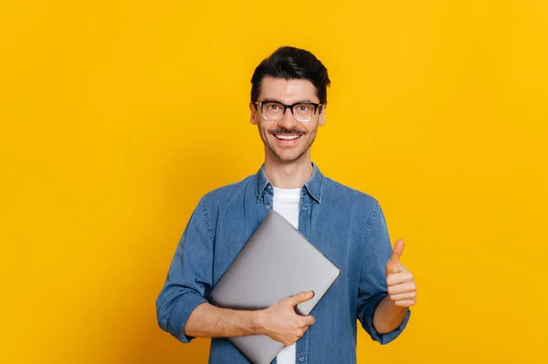 Attractive smart caucasian guy, with glasses, student or freelancer, holding a laptop in hand, standing against isolated orange background, looking at camera, smiling friendly, shows thumb up gesture — Stockfoto