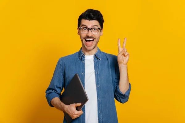 Friendly positive caucasian stylish handsome guy with glasses, freelancer or student, holds a laptop in hand, shows a peaceful gesture, stands on isolated orange background, looks at camera, smiling — Stockfoto