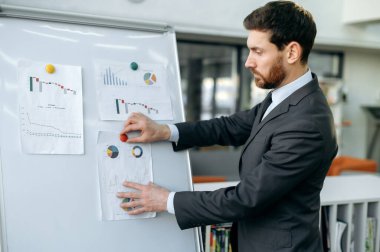 Pensive intelligent Caucasian adult businessman, financial analyst, department head, stands near the whiteboard in office, analyzes financial charts, ponders financial risks, profit, business strategy