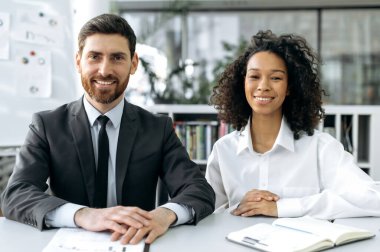Portrait of two successful multiracial business people. Caucasian successful business man in a suit, and African American pretty woman in white shirt, in modern office, looking at the camera, smile