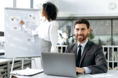 Intelligent adult Caucasian businessman, in suit, sits at a table in modern office, uses laptop, looks into the camera, smiles. On the background a female colleague studies charts on a whiteboard